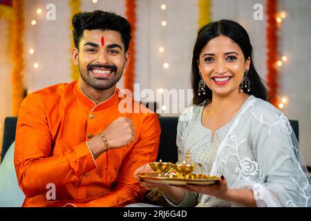 Heureux frère indien souriant montrant rakhi avec sa sœur en regardant la caméra à la maison - concept de relation familiale, cérémonie de tradition et indien Banque D'Images