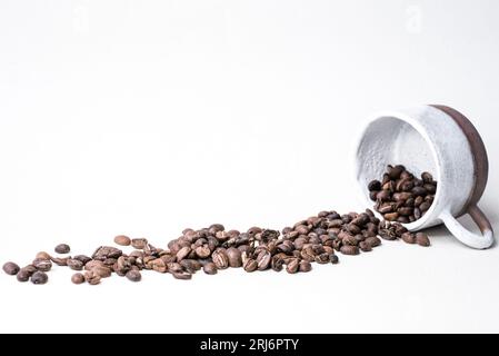 Un tas de grains de café fraîchement torréfiés se déversant d'une tasse en céramique blanche sur un fond blanc solide Banque D'Images