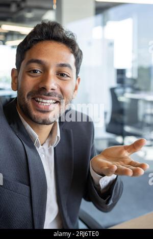 Photo verticale, jeune homme d'affaires prospère regardant dans la caméra smartphone parler sur appel vidéo en utilisant l'application sur le téléphone, souriant et ayant une conversation amicale avec des amis et des collègues à l'intérieur du bureau. Banque D'Images