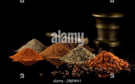 Tas d'épices multicolores - origan, tacos, graines de lin, poivre noir et rouge, clous de girofle et mortier en bronze sur fond miroir noir Banque D'Images
