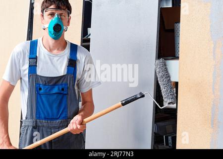 C'est peintre professionnel dans le respirateur et des lunettes avec rouleau à peinture dans ses mains. Portrait d'un véritable ouvrier. Peintre regarde dans la caméra. Banque D'Images