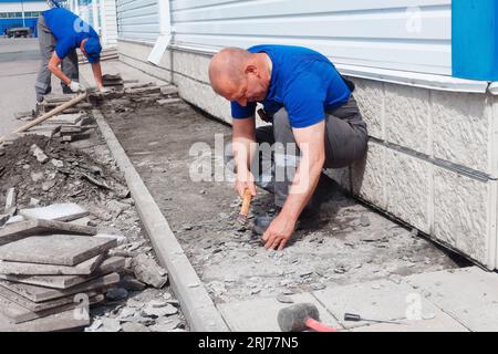 Le maçon âgé pose des tuiles sur la fondation du bâtiment à l'extérieur. Entrepreneur en construction pose des tuiles de parement de façade de l'installation de production. L'homme pose des carreaux le jour d'été. Banque D'Images