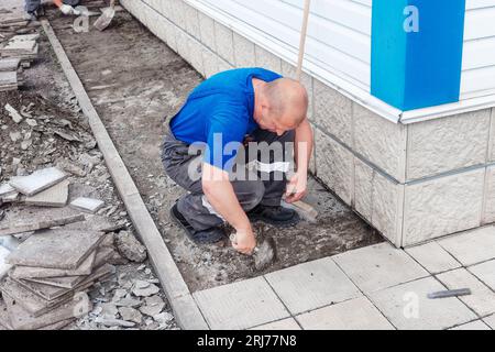 Le maçon âgé pose des tuiles sur la fondation du bâtiment à l'extérieur. Entrepreneur en construction pose des tuiles de parement de façade de l'installation de production. L'homme pose des carreaux le jour d'été. Banque D'Images