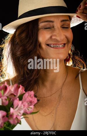 Studio portrait d'une belle femme mûre portant un chapeau tenant un bouquet de fleurs entre les mains. Isolé sur fond noir. Banque D'Images