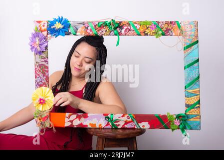 Photo de belle femme avec des tresses dans les cheveux derrière un cadre décoré de fleurs et de tissus colorés dans une position horizontale touchant la fleur Banque D'Images