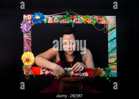 Photo de belle femme avec des tresses dans les cheveux à l'intérieur d'un cadre orné de fleurs et de tissus colorés dans une position horizontale touchant les fleurs Banque D'Images