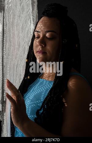 Photo d'une belle femme avec des tresses dans les cheveux et des ombres géométriques sur son visage avec ses mains sur un tissu blanc. Portrait de studio avec fond noir Banque D'Images