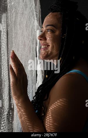 Photo d'une belle femme avec des tresses dans les cheveux et des ombres géométriques sur son visage avec ses mains sur un tissu blanc. Portrait de studio avec fond noir Banque D'Images