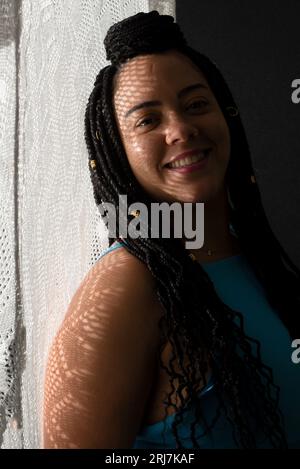 Photo d'une jolie femme avec des tresses dans les cheveux et des ombres géométriques sur son visage appuyé contre un tissu blanc. Portrait de studio avec fond noir Banque D'Images