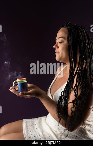 Portrait d'une belle femme avec des tresses dans les cheveux, assise, portant des vêtements blancs avec deux tasses de café à la vapeur. Studio photographié avec une lila sombre Banque D'Images