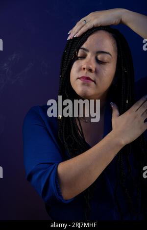 Une belle femme aux cheveux tressés en vêtements bleus, touchant la tête et la poitrine. souriant et heureux. Isolé sur fond bleu foncé. Perso positif Banque D'Images