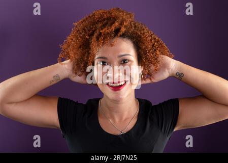 une femme aux cheveux roux séduisante posant pour une photo en vêtements noirs touchant ses cheveux. Confiant et heureux. Isolé sur fond de couleur lilas. Banque D'Images