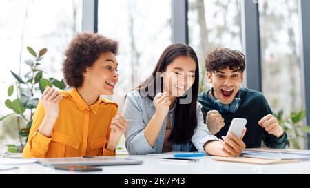 Étudiants multiraciaux excités regardant le téléphone portable et serrant les poings, vérifiant les résultats des examens en ligne dans la salle de classe Banque D'Images