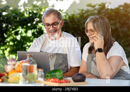 Sourire vieux mari et femme caucasiens dans des tabliers utilisent un ordinateur portable, font des appels téléphoniques à table avec des légumes biologiques Banque D'Images