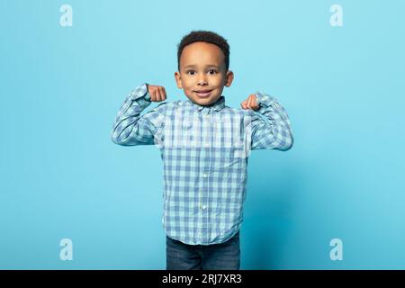 Drôle petit garçon noir montrant des biceps et souriant à la caméra, enfant mâle démontrant sa puissance et sa force Banque D'Images