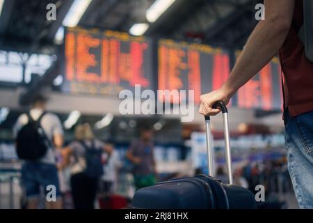 Voyager en avion. Homme attendant dans le terminal de l'aéroport. Focalisation sélective sur la main tenant la valise contre le tableau d'arrivée et de départ. Le passager est rea Banque D'Images