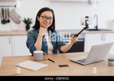 Femme énergique se filmant sur selfie bâton avec smartphone moderne pour blog personnel. Femme asiatique positive dans les lunettes enregistrant des cours en ligne wh Banque D'Images