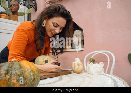femme caucasienne à la maison debout, heureuse, souriante et dessinant citrouille halloween avec marqueur noir, concept de vacances et traditions américaines Banque D'Images