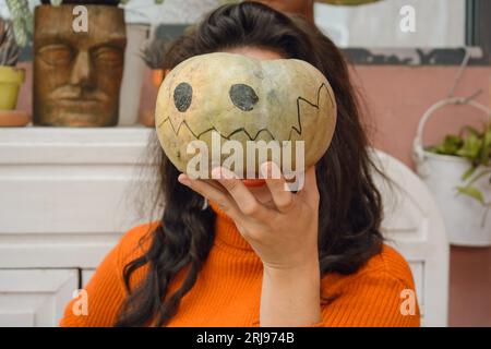 femme caucasienne adulte dans des vêtements orange, méconnaissable avec de longs cheveux, debout à la maison couvrant son visage avec la citrouille d'halloween, concept de gens, flic Banque D'Images