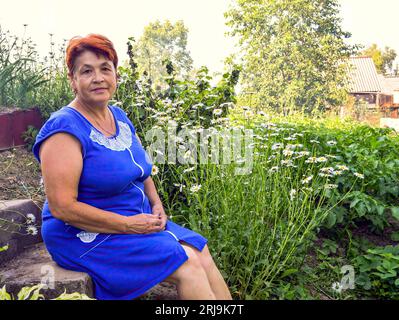 Portrait d'une femme adulte des années 60 en robe bleue assise à côté pour fleurir des lits avec des pâquerettes dans le village en été soirée Banque D'Images