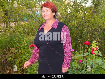 Vieille belle femme en robe souriante et debout près de beaux asters roses et rouges. Banque D'Images