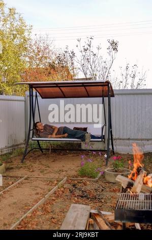 Jeune homme endormi allongé sur une balançoire de jardin à côté d'un feu de joie Banque D'Images