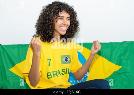 Fan de football féminin brésilien acclamé avec drapeau du Brésil regardant le match à la télévision à l'intérieur à la maison Banque D'Images