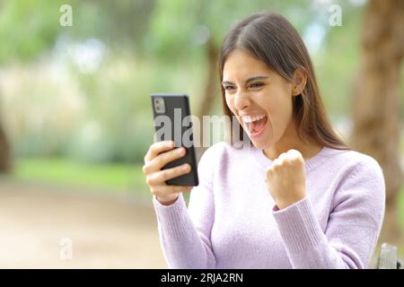 Femme excitée célébrant les bonnes nouvelles regardant le contenu du téléphone dans un parc Banque D'Images