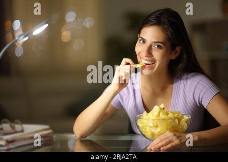 Femme heureuse dans la nuit mangeant des chips de pomme de terre regardant la caméra à la maison Banque D'Images