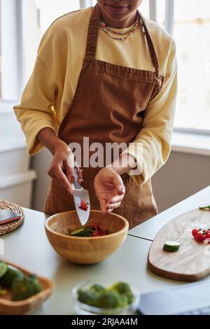 Portrait vertical de jeune femme noire ajoutant des légumes à un bol en bois tout en cuisinant la salade dans la cuisine Banque D'Images