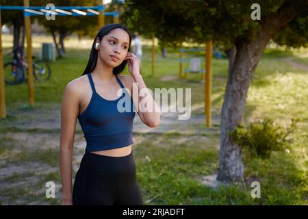 Jeune femme biraciale en vêtements de sport écoutant de la musique sur des écouteurs sans fil dans le parc Banque D'Images