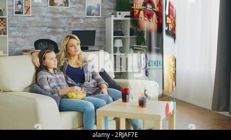 Famille heureuse choisissant quelle émission de télévision regarder sur l'écran de projecteur AR. Mère et fille se détendant à la maison sur un canapé, regardant du contenu en ligne sur des services de streaming tout en dégustant des collations et des boissons. Banque D'Images