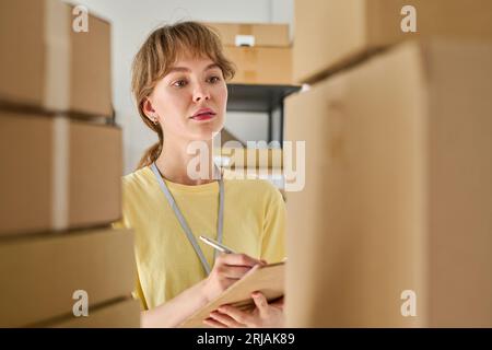 Jeune femme sérieuse en t-shirt jaune prenant des notes dans le document tout en regardant la pile de boîtes en carton emballées debout sur l'étagère Banque D'Images