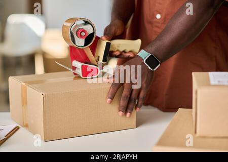Mains de jeune homme afro-américain travailleur de marché ou de salle de stockage préparant des colis avec des marchandises en ligne pour l'envoi aux clients Banque D'Images