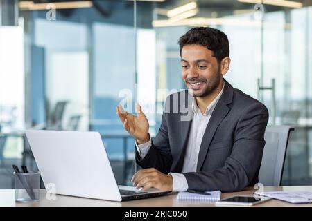 Jeune homme d'affaires arabe prospère dans le bureau au travail parlant à distance avec des collègues et des partenaires, homme utilisant un ordinateur portable pour les appels vidéo, patron satisfait souriant et faisant des gestes avec les mains joyeusement. Banque D'Images