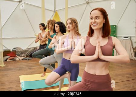 femme heureuse regardant loin, debout avec des mains priantes pendant le cours de yoga avec des copines multiethniques Banque D'Images