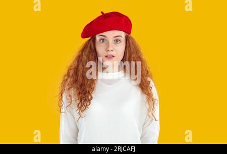 Fille rousse souriante effrayée en béret rouge et chandail blanc regardant la caméra sur fond jaune. Banque D'Images