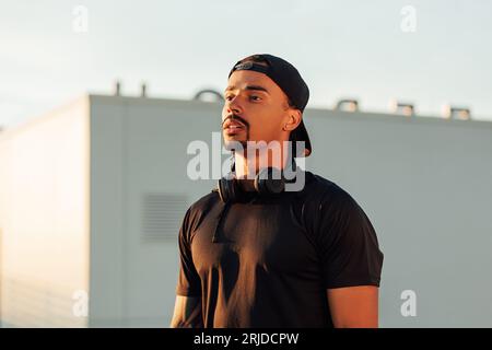 Portrait d'un jeune athlète masculin confiant dans une casquette. Athlète avec des écouteurs sans fil debout sur un toit au coucher du soleil. Banque D'Images