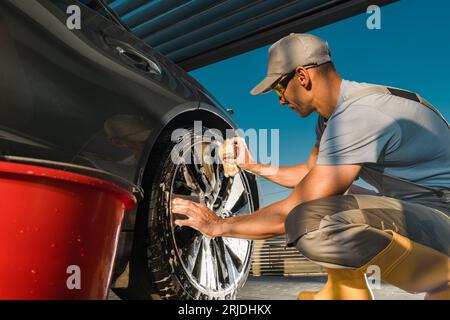 Travailleur caucasien nettoyant des roues en alliage de véhicule moderne en utilisant Pro détergent à l'intérieur d'un abri d'auto Banque D'Images