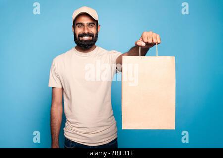 Heureux livreur indien en uniforme tenant et montrant le sac en papier artisanal à la caméra, fond bleu, espace pour la conception Banque D'Images