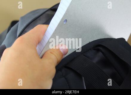 Une photo d'un étudiant blanc mettant son carnet de papier dans son sac d'école gris. Banque D'Images