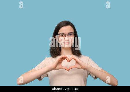 Belle étudiante fille en lunettes fait une forme de coeur avec ses mains, sourit sur fond bleu. Amour, relations, Saint-Valentin. Banque D'Images