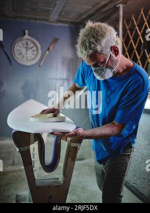 Vue de côté du maître masculin blond concentré dans le masque de respirateur polir la planche de surf blanche placée sur des supports de façonnage dans l'atelier Banque D'Images