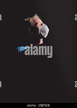 Vue latérale d'un homme blond coiffé d'un masque respiratoire protecteur et les yeux fermés dans l'obscurité sur fond noir éclairé par la lumière du soleil Banque D'Images