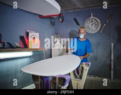 Shaper masculin blond concentré dans le masque respiratoire examinant la forme de la planche placée sur des supports tout en créant des planches de surf à la main dans l'atelier Banque D'Images