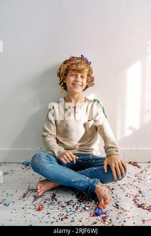 Garçon préadolescent joyeux dans des vêtements décontractés assis avec les jambes croisées sur le plancher en bois recouvert de confettis et d'étincelles Banque D'Images