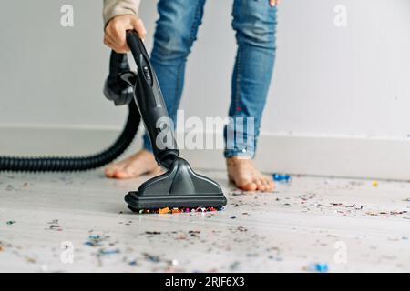 Crop anonyme garçon pieds nus debout sur parquet recouvert de confettis dispersés et en utilisant un aspirateur moderne Banque D'Images