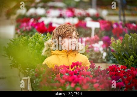 Garçon aux cheveux bouclés preteen rêveur assis sur un banc près de fleurs colorées en fleurs et regardant loin Banque D'Images