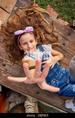 Vue de dessus de fille joyeuse en denim dans l'ensemble regardant la caméra avec le bras levé tout en se reposant sur un banc en bois dans la campagne Banque D'Images