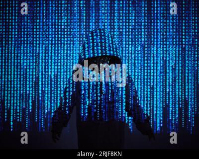 Garçon hacker anonyme en sweat à capuche debout dans la projection du code binaire bleu dans la chambre noire Banque D'Images
